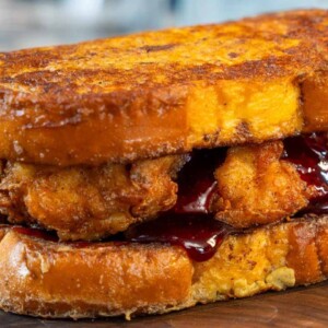 A close-up of a fried chicken sandwich with thick, golden-brown bread slices, crispy fried chicken, and dark red sauce, resting on a wooden surface.