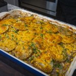 A classic beef and potato casserole, topped with golden-brown sliced potatoes and sprinkled with chopped herbs, sits in a rectangular dish on a dark wooden surface in a cozy kitchen setting. Perfect for fans of ground beef and potato casserole recipes.