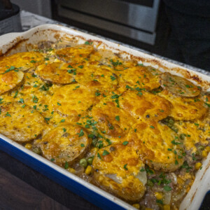 A classic beef and potato casserole, topped with golden-brown sliced potatoes and sprinkled with chopped herbs, sits in a rectangular dish on a dark wooden surface in a cozy kitchen setting. Perfect for fans of ground beef and potato casserole recipes.