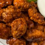 Close-up of saucy, crispy boneless wings piled on a plate with a small cup of creamy white dipping sauce on the side. Perfect for trying your favorite boneless wings recipe at home!.