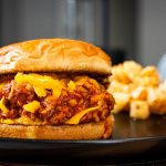 A chicken sloppy joe sandwich with shredded cheddar cheese on a toasted bun sits on a black plate, with a serving of crinkle-cut fries in the background.