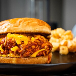 A chicken sloppy joe sandwich with shredded cheddar cheese on a toasted bun sits on a black plate, with a serving of crinkle-cut fries in the background.