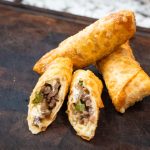 Four golden-brown egg rolls rest on a dark wooden surface, with one egg roll cut open to reveal a filling of ground beef and green peppers—an irresistible glimpse into this savory egg roll recipe.