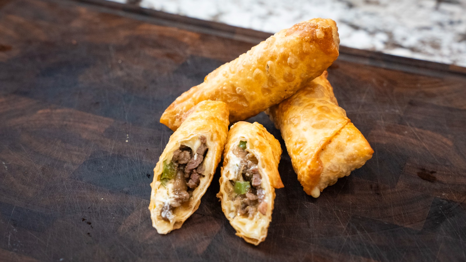 Four golden-brown egg rolls rest on a dark wooden surface, with one egg roll cut open to reveal a filling of ground beef and green peppers—an irresistible glimpse into this savory egg roll recipe.