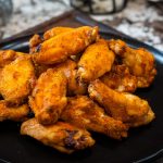 A black plate piled with golden-brown, seasoned air fryer chicken wings sits on a dark surface, with a blurred background.