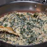 A large skillet filled with creamy spinach and mushrooms, perfect for appetizer dips like collard green dip, is being stirred with a wooden spatula on a granite countertop.