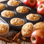 Freshly baked apple cinnamon muffins in a muffin tray and paper liners, surrounded by red apples and cinnamon sticks on a rustic wooden table.