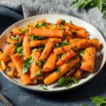 A plate of roasted carrots, cut into sticks and garnished with chopped fresh parsley, sits on a dark tabletop beside a silver spoon and a gray napkin.