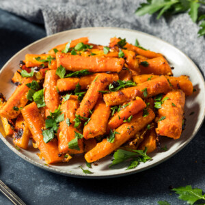 A plate of roasted carrots, cut into sticks and garnished with chopped fresh parsley, sits on a dark tabletop beside a silver spoon and a gray napkin.