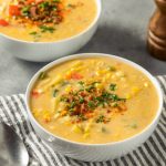 Two bowls of creamy corn chowder, garnished with chopped herbs, sit on a striped cloth with two metal spoons beside them. A pepper grinder and part of a white item are visible in the background.