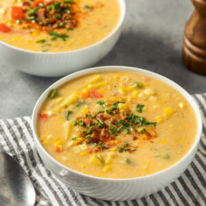 Two bowls of creamy corn chowder, garnished with chopped herbs, sit on a striped cloth with two metal spoons beside them. A pepper grinder and part of a white item are visible in the background.