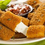Plate of golden-brown breaded mozzarella sticks with melted cheese inside, served with a small bowl of marinara sauce in the background and garnished with herbs—perfectly crispy mozzarella sticks for snacking.