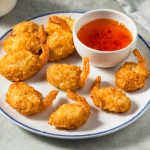 A plate of crispy coconut shrimp is arranged around a small bowl of orange coconut shrimp sauce, set on a light-colored surface with a striped cloth partially visible in the background.