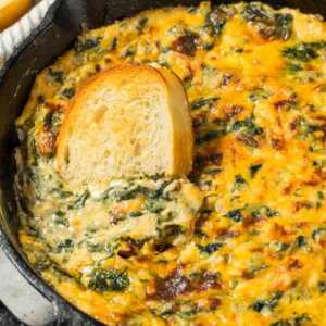 A skillet filled with creamy, baked spinach dip, with a slice of toasted bread partially dipped into the mixture.