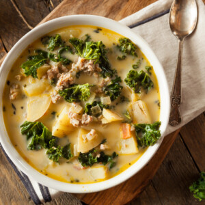 A bowl of creamy Tuscan Soup with kale, potato chunks, and sausage sits on a wooden table next to a spoon and napkin. The soup is hearty and rustic, garnished with fresh green kale leaves.