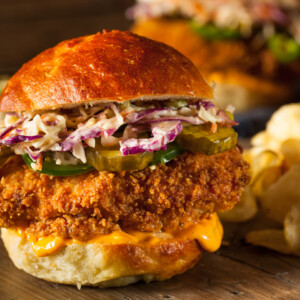 A spicy chicken sandwich with pickles, shredded coleslaw, and orange sauce on a brioche bun, served on a wooden surface with potato chips on the side.