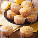 A plate of lemon muffins with white icing and lemon zest sits on a rustic wooden table, with more cupcakes and lemon slices nearby on a yellow-striped cloth.