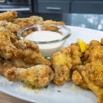 A platter of golden fried garlic parmesan wings is served with a bowl of creamy dipping sauce, garnished with herbs and lemon wedges, on a white plate in a kitchen setting.
