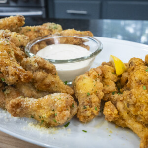 A platter of golden fried garlic parmesan wings is served with a bowl of creamy dipping sauce, garnished with herbs and lemon wedges, on a white plate in a kitchen setting.