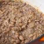 A close-up of cooked pinto beans mixed with shredded meat and herbs in a large orange pot shows a delicious pinto beans recipe, with a wooden spoon partially visible stirring the mixture.