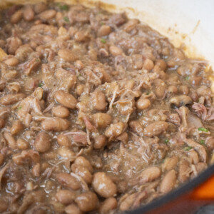 A close-up of cooked pinto beans mixed with shredded meat and herbs in a large orange pot shows a delicious pinto beans recipe, with a wooden spoon partially visible stirring the mixture.