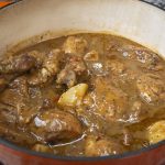 A close-up of a red Dutch oven filled with a mouthwatering chicken stew recipe—braised chicken, potatoes, and vegetables simmer in a rich, brown, seasoned sauce on the stovetop.