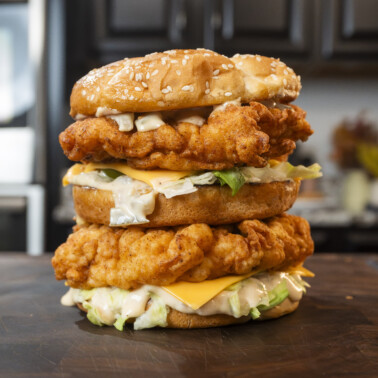 A towering McDonald's Chicken Big Mac sandwich with crispy fried chicken fillets, lettuce, cheese, and sauce between three sesame seed buns, placed on a wooden surface in a kitchen setting.