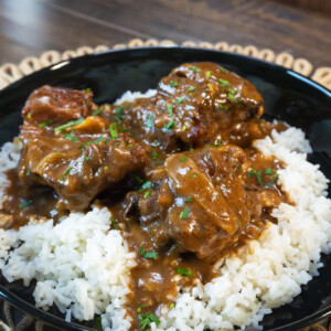 A black bowl filled with white rice topped with savory, brown gravy-covered Slow Cooker Oxtails, garnished with chopped herbs, sits on a woven placemat on a wooden table.