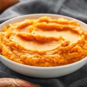 A white bowl filled with creamy mashed sweet potatoes sits on a dark cloth, showcasing an inviting mashed sweet potatoes recipe with whole sweet potatoes and green herbs in the background.