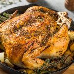 A golden-brown roasted whole chicken in oven, seasoned with herbs, sits in a cast iron skillet, surrounded by roasted potatoes and onions, garnished with fresh thyme sprigs.