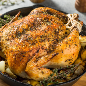 A golden-brown roasted whole chicken in oven, seasoned with herbs, sits in a cast iron skillet, surrounded by roasted potatoes and onions, garnished with fresh thyme sprigs.
