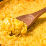 A close-up of a baked corn bread casserole in a skillet, with a wooden spoon scooping out a portion, showing its creamy and golden texture.