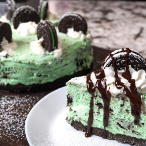 A slice of mint Oreo cheesecake is topped with whipped cream, chocolate sauce, and a cookie, with the rest of this easy cheesecake recipe and extra cookies in the background on a powdered sugar-dusted surface.