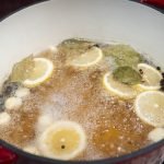 A red pot on a stove contains a bubbling chicken brine recipe with lemon slices, garlic cloves, bay leaves, and peppercorns—perfect for anyone wondering how to brine chicken.
