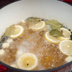 A red pot on a stove contains a bubbling chicken brine recipe with lemon slices, garlic cloves, bay leaves, and peppercorns&mdash;perfect for anyone wondering how to brine chicken.