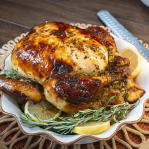 A roasted whole chicken, prepared using a classic roast chicken recipe, is garnished with fresh rosemary and lemon slices on a white plate atop a rustic wooden table, with a large knife resting nearby.