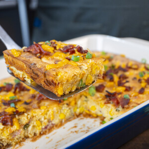A close-up of a spatula lifting a square piece of cheesy baked cornbread casserole topped with bacon and green onions from a dish. The golden cornbread casserole recipe features visible corn kernels for extra flavor.