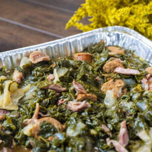 A close-up of a tray filled with savory collard greens mixed with pieces of meat, likely ham or sausage, on a wooden table with yellow flowers in the background&mdash;perfect for trying out your favorite collard greens recipe.