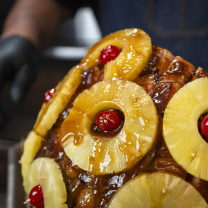 Close-up of a bourbon glazed ham decorated with pineapple rings and maraschino cherries; a person wearing black gloves is seen in the background, slightly out of focus.