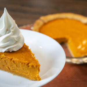 A slice of pumpkin pie topped with whipped cream on a white plate, with the remaining pie in a glass dish in the background, missing a slice—perfect inspiration if you're searching for the best sweet potato pie recipe.