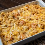 A white casserole dish filled with seafood mac and cheese, topped with melted cheese and shrimp, rests on a wooden table with a woven placemat underneath.