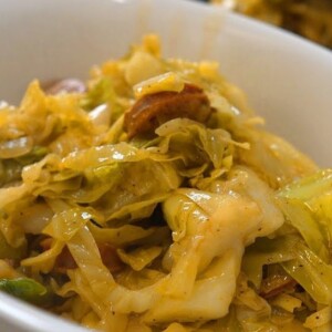 A close-up of a white bowl filled with savory fried cabbage and sausage slices, seasoned and mixed together, with a fork partially visible&mdash;perfect for anyone craving a classic fried cabbage recipe.