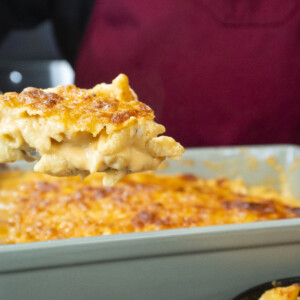 A close-up of a spoonful of creamy baked mac and cheese being lifted from a casserole dish, with a golden, crispy topping and a person in a red apron in the background.