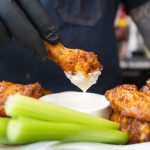 A person wearing black gloves dips a saucy buffalo wing into a cup of white dipping sauce, surrounded by more wings and celery sticks—a perfect scene for showcasing your favorite buffalo wings recipe.