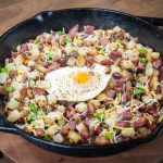 A cast iron skillet filled with corned beef hash—diced potatoes, corned beef, melted cheese, and green onions—topped with a sunny side up egg, sits on a wooden table. Perfect for trying out new corned beef recipes.