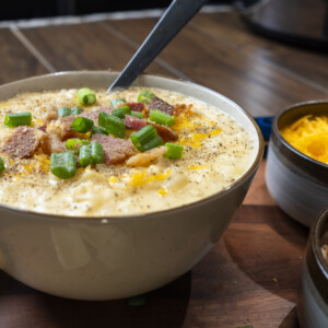 A bowl of creamy loaded potato soup topped with chopped green onions, bacon bits, and black pepper. Nearby are small bowls of shredded cheese and more bacon, with a chunk of cheese beside the bowl on a wooden surface.