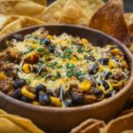 A bowl of chili topped with shredded cheese and herbs, filled with black beans, corn, and ground meat, surrounded by crispy tortilla chips—a comforting dish inspired by classic taco casserole recipes.
