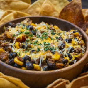 A bowl of chili topped with shredded cheese and herbs, filled with black beans, corn, and ground meat, surrounded by crispy tortilla chips&mdash;a comforting dish inspired by classic taco casserole recipes.