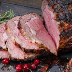 Sliced prime rib roast on a cutting board, garnished with fresh herbs and cranberries, set against a rustic wooden background.