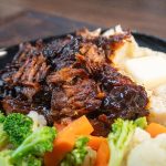 A plate of crock pot bbq beef roast with dark sauce, mashed potatoes topped with a pat of butter, and a side of steamed broccoli, carrots, and cauliflower.